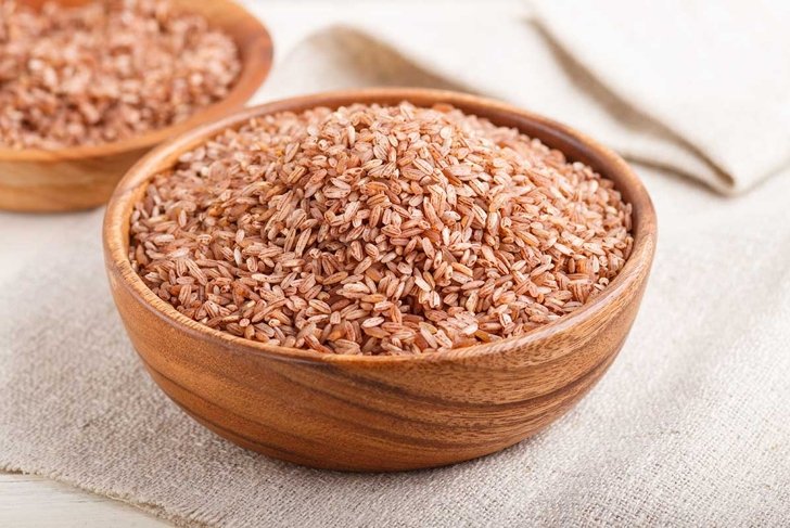 Two wooden bowls with unpolished brown rice on a white wooden background and linen textile. Side view, close up, selective focus.