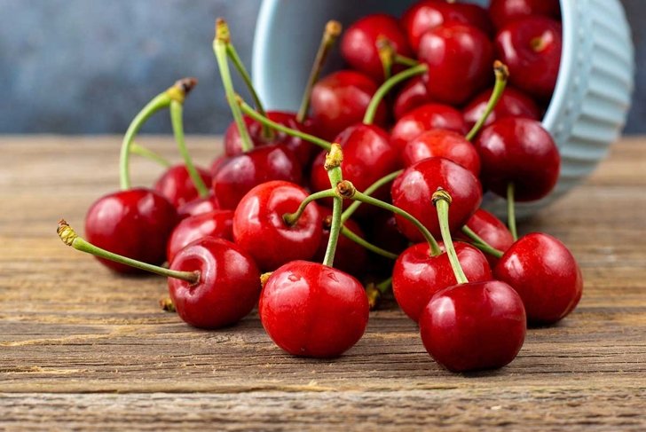 Ripe wet sweet cherries are poured out of the blue bowl on wooden background