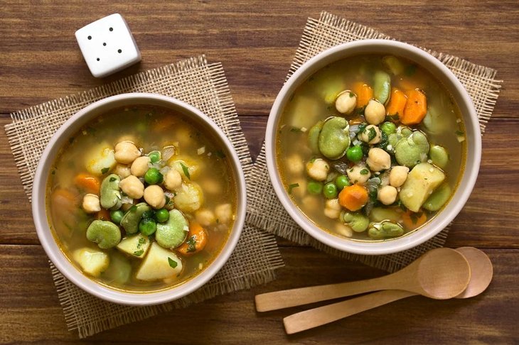 Vegetarian chickpea soup with carrot, broad bean (fava bean), pea, potato, onion, garlic and parsley served in bowls, photographed overhead on wood with natural light